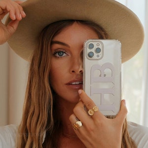 Könnte beinhalten: Eine transparente Handyhülle mit den Buchstaben "HB" in hellem Lila. Die Handyhülle wird vor dem Gesicht einer Person gehalten. Die Person trägt einen beigen Hut, ein weißes Hemd und goldene Ringe.