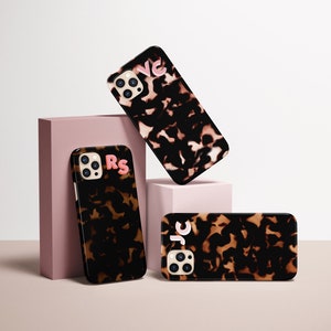 May include: Three smartphone cases with a tortoiseshell pattern and personalized initials. The cases are black and brown with pink and white letters. One case is upright, one is angled, and one is flat.