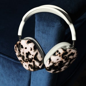 May include: Over-ear headphones with a white headband and ear cushions. The earcups have a tortoiseshell pattern in shades of pink, black, and brown. The headphones are resting on a blue velvet surface.