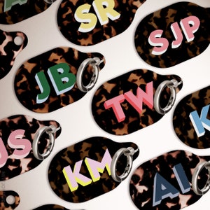 May include: A selection of oval keyrings with a tortoiseshell design. Each keyring displays a different set of initials in bold, colourful letters, and a silver hoop earring. The initials include: SR, SJP, JB, TW, KM, and AI.