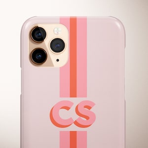 May include: A light pink phone case with a vertical stripe design in shades of pink and orange. The case features the initials "CS" in a bold, layered font. The phone's camera lenses are visible at the top.