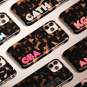 May include: A collection of smartphone cases with a tortoiseshell pattern. Each case features a different name or initials in a bold, colorful font. The cases are arranged on a light pink surface.