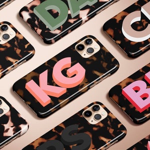 May include: A collection of smartphone cases with a tortoiseshell pattern. Each case features a different set of 3D letters in various colors, including pink, green, and blue. The cases are arranged on a light brown surface.