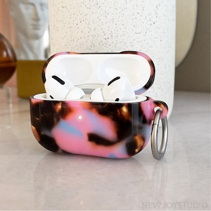 May include: A pair of white wireless earbuds inside a patterned case. The case has a pink, brown, and blue tortoiseshell design with a silver-colored ring. The case is open, revealing the earbuds.