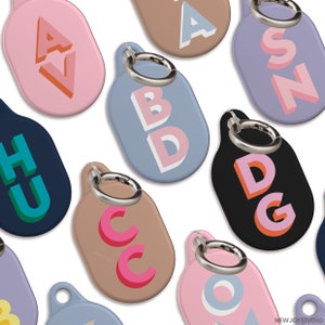 Peut inclure: Plusieurs porte-clés ovales colorés avec des anneaux argentés. Chaque porte-clé présente une combinaison de lettres différente dans un style 3D audacieux. Les porte-clés sont de différentes couleurs, dont le rose, le bleu, le marron et le noir.