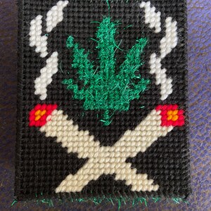 May include: A black canvas with a green marijuana leaf and two white cigarettes with red and orange flowers at the ends. The cigarettes are crossed over each other.
