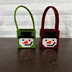 May include: Two small, handmade snowman-themed bags. One is green with a black hat and handle, the other is burgundy with a black hat and handle. Both have white faces with black eyes and smiles, and orange carrot noses. The bags are on a brown surface.