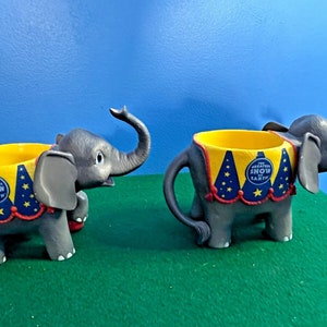 Puede incluir: Dos tazas de plástico en forma de elefante gris con diseños de carpa de circo de color amarillo, rojo, azul y blanco. Las tazas tienen el texto "The Greatest Show on Earth".