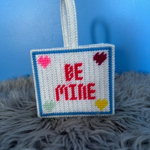 May include: A white, square, needlepoint ornament with the words "BE MINE" in red. The ornament has a blue border and features pink, maroon, yellow, and green heart-shaped accents. A white ribbon is attached for hanging.