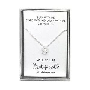 May include: A silver necklace with a small, clear crystal pendant in a white box. The box has the text "PLAN WITH ME STAND WITH ME LAUGH WITH ME CRY WITH ME WILL YOU BE Bridesmaid? doodlebeads.com" printed on it.