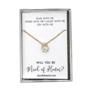 May include: A small white box with a gold necklace inside. The necklace has a small, clear stone pendant. The box has the text "PLAN WITH ME STAND WITH ME LAUGH WITH ME CRY WITH ME WILL YOU BE Maid of Honor? doodlebeads.com" printed on it.