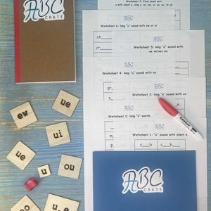 May include: Educational materials for learning. The image shows a brown notebook, worksheets, wooden letter tiles, and a blue folder, all with the "ABC CRATE" logo. A red marker is also present. The worksheets focus on phonics.
