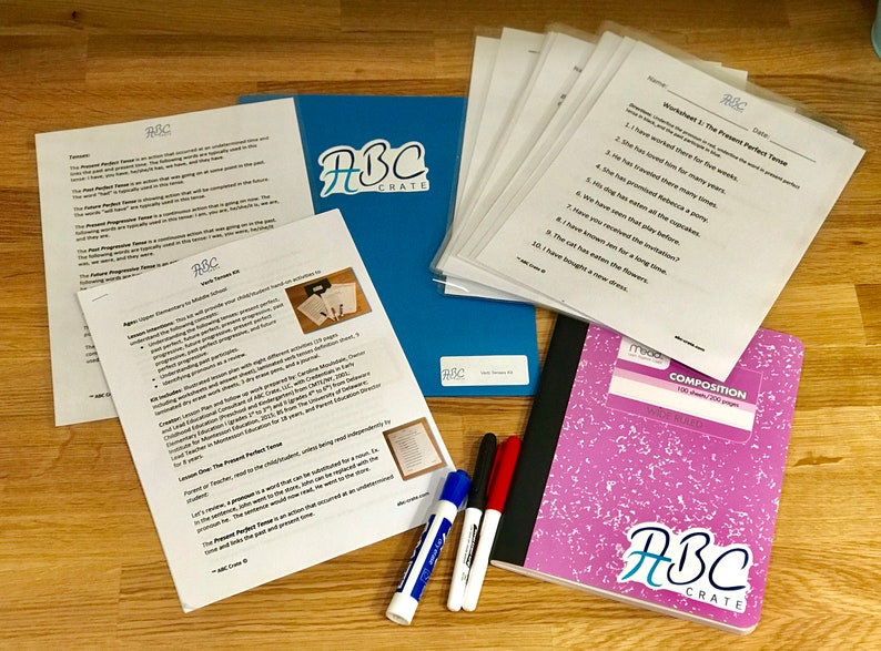 Puede incluir: Un cuaderno de composici&oacute;n rosa con el logotipo de ABC Crate en la portada, junto con una carpeta azul con el logotipo de ABC Crate, y varias hojas blancas con el t&iacute;tulo "Worksheet 1: The Present Perfect Tense". Tambi&eacute;n hay tres marcadores de borrado en seco blancos y un marcador de borrado en seco rojo.