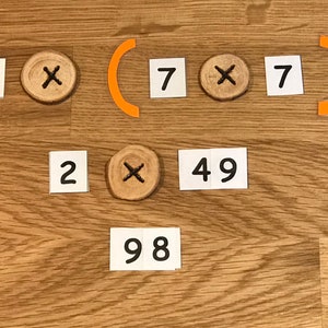 Puede incluir: Se muestra un problema de matem&aacute;ticas con la ecuaci&oacute;n 2 x (7 x 7) = 98. La ecuaci&oacute;n se resuelve usando tarjetas blancas con n&uacute;meros y un c&iacute;rculo de madera con un s&iacute;mbolo de multiplicaci&oacute;n.