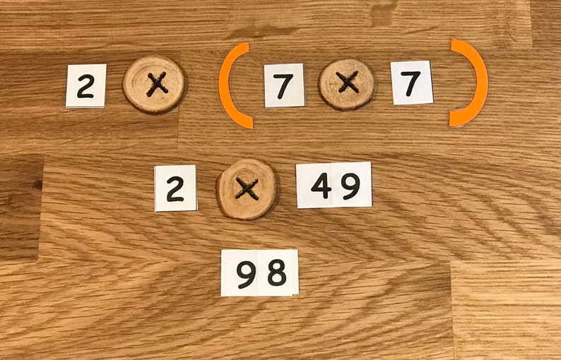 Puede incluir: Se muestra un problema de matem&aacute;ticas con la ecuaci&oacute;n 2 x (7 x 7) = 98. La ecuaci&oacute;n se resuelve usando tarjetas blancas con n&uacute;meros y un c&iacute;rculo de madera con un s&iacute;mbolo de multiplicaci&oacute;n.
