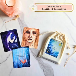 Peut inclure: Un ensemble de trois cartes carrées avec des illustrations à l'aquarelle et du texte, incluant un portrait, une silhouette dans l'eau bleue profonde et une forme abstraite sombre, avec une pochette à cordon crème étiquetée Emotional Witness.