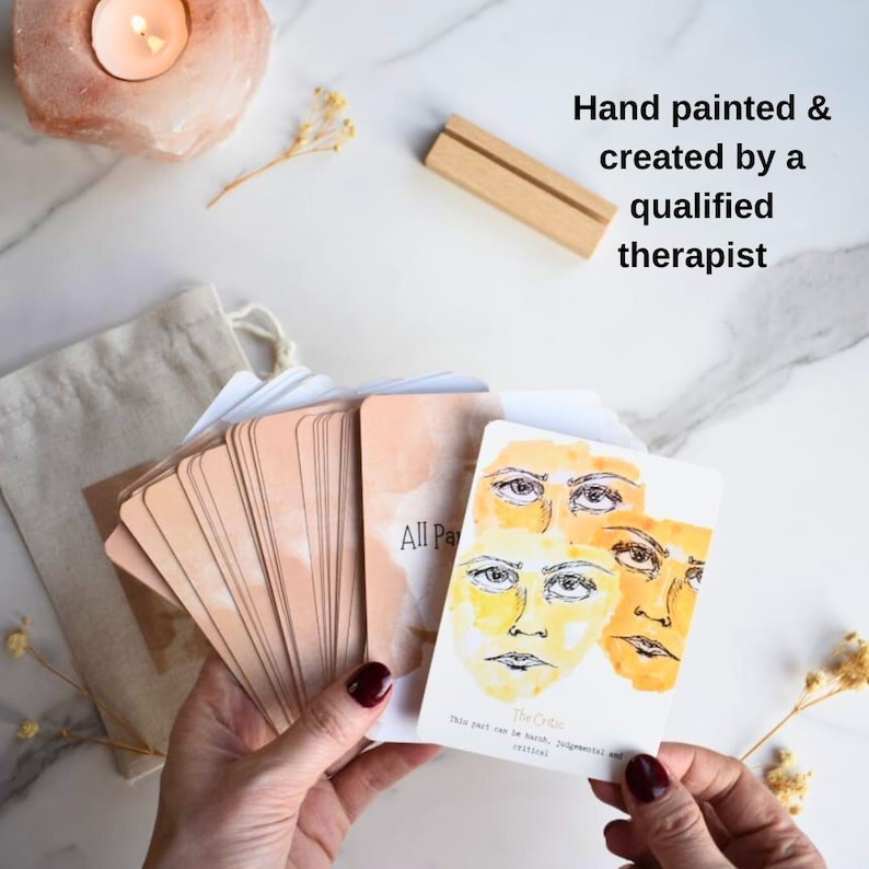 May include: A deck of rectangular cards with watercolor illustrations of human faces in shades of yellow and orange. The top card features three faces and the text 'The Critic' with a description. Includes a small wooden stand and a fabric pouch.