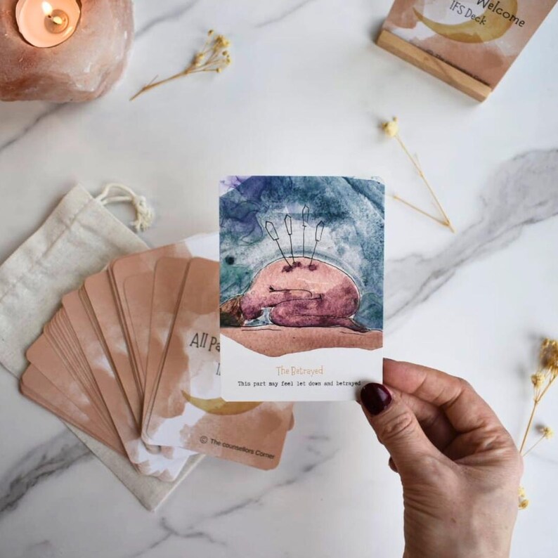 May include: A rectangular card from a deck featuring a watercolor illustration of a figure curled in a fetal position with three swords piercing its back. The text reads The Betrayed and This part may feel let down and betrayed.