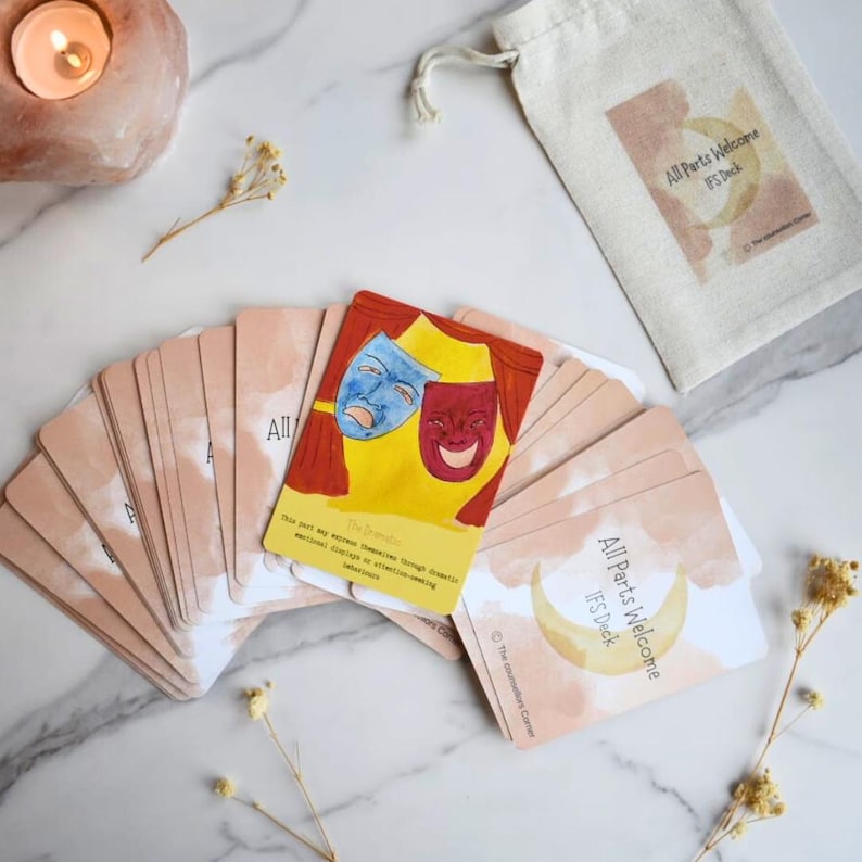 May include: A deck of All Parts Welcome IFS cards with a beige drawstring bag. One card features a yellow background with red curtains and blue and red theatrical masks. Other cards show a crescent moon and the text All Parts Welcome IFS Deck.