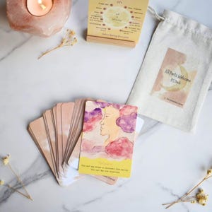 May include: A deck of cards featuring watercolor illustrations of faces and clouds, a yellow instruction card titled Unburdening Process, and a beige drawstring pouch labeled All Parts Welcome IFS Deck, arranged on a white marble surface.