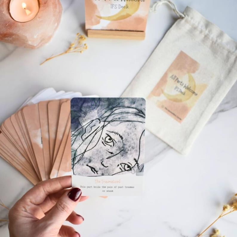 May include: A deck of cards titled All Parts Welcome IFS Deck, featuring a card with a black and white line drawing of a face and the text The Traumatized. This part holds the pain of past traumas or shame, alongside a matching cotton drawstring bag.