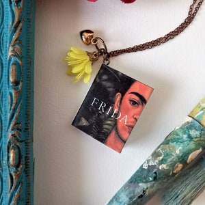 Pu&ograve; includere: Una collana con catena dorata con un piccolo ciondolo a forma di libro con un ritratto di Frida Kahlo. La copertina del libro dice "Frida" e ha un ciondolo di fiore giallo attaccato.