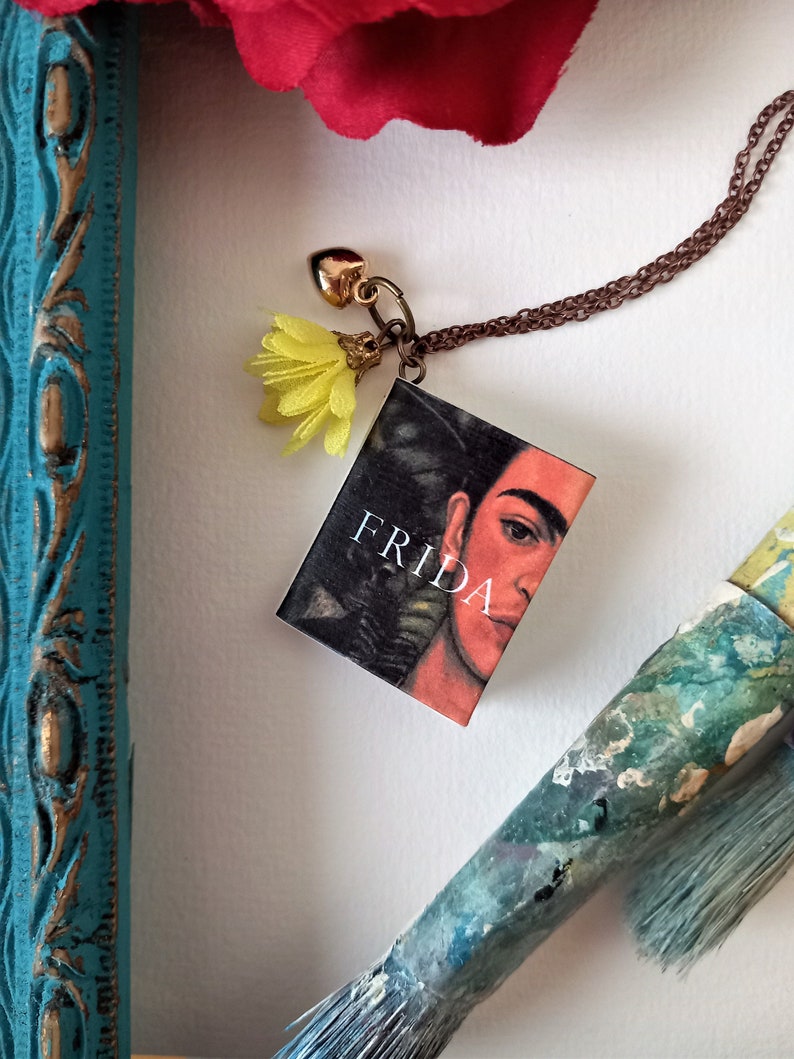 Pu&ograve; includere: Una collana con catena dorata con un piccolo ciondolo a forma di libro con un ritratto di Frida Kahlo. La copertina del libro dice "Frida" e ha un ciondolo di fiore giallo attaccato.