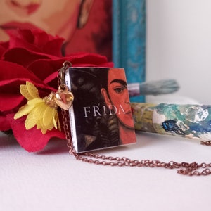 Pu&ograve; includere: Una collana con un ciondolo a forma di cuore dorato e un piccolo libro con un'immagine in bianco e nero di Frida Kahlo e la parola "FRIDA" stampata sulla copertina.