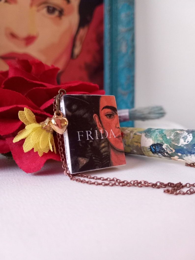 Pu&ograve; includere: Una collana con un ciondolo a forma di cuore dorato e un piccolo libro con un'immagine in bianco e nero di Frida Kahlo e la parola "FRIDA" stampata sulla copertina.