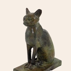 Puede incluir: Una estatua de bronce de un gato sentado. El gato está representado en un estilo realista con detalles precisos, incluyendo su pelaje, ojos y patas. La estatua está montada sobre una base rectangular.