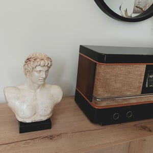 K&ouml;nnte beinhalten: Eine cremefarbene B&uuml;ste eines Mannes mit Lorbeerkranz, auf einem schwarzen Sockel. Daneben ein Vintage-Radio in Schwarz mit gewebter Front und braunen Holzakzenten. Die Gegenst&auml;nde stehen auf einer Holzoberfl&auml;che.