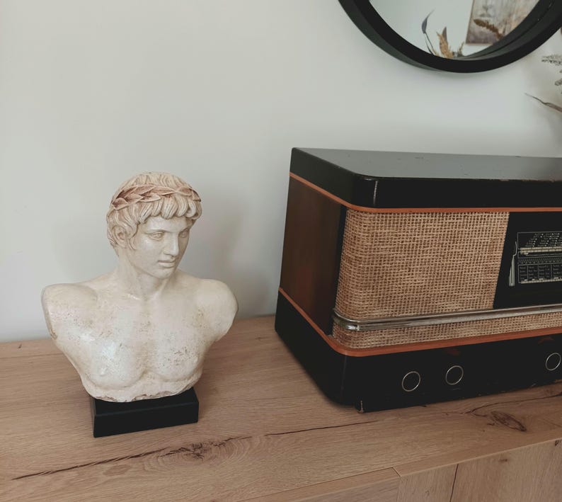 K&ouml;nnte beinhalten: Eine cremefarbene B&uuml;ste eines Mannes mit Lorbeerkranz, auf einem schwarzen Sockel. Daneben ein Vintage-Radio in Schwarz mit gewebter Front und braunen Holzakzenten. Die Gegenst&auml;nde stehen auf einer Holzoberfl&auml;che.