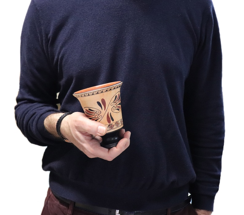 May include: A small, ceramic cup with a brown and red painted design. The cup has a wide, round base and a narrow, flared top. The design features a stylized floral pattern with a geometric border.