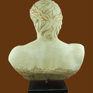 K&ouml;nnte beinhalten: Cremefarbene B&uuml;ste einer Person, von hinten gesehen, mit Lorbeerkranz. Die Skulptur ist auf einem schwarzen, rechteckigen Sockel vor braunem Hintergrund montiert. Die B&uuml;ste hat detaillierte Haare und Schulterdefinition.