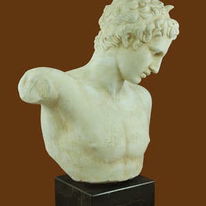 Puede incluir: Un busto de yeso blanco de un hombre joven con el pelo corto y rizado. El busto est&aacute; sobre una base de m&aacute;rmol negro.