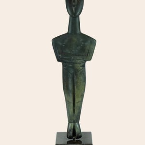 May include: A green bronze sculpture of a standing figure with a simplified, geometric design. The figure has a long neck, a small head, and arms crossed at the chest. The sculpture is mounted on a black marble base.
