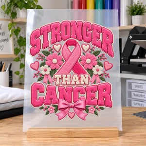 May include: A clear acrylic sign featuring a pink ribbon graphic with the text STRONGER THAN CANCER in bold pink letters. The design includes pink flowers, small white blossoms, green leaves, and a pink bow, all mounted on a wooden base.