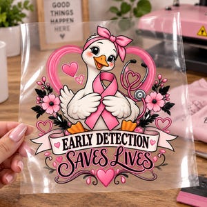 May include: A clear heat transfer sheet featuring a cartoon goose wearing a pink headband and holding a pink awareness ribbon. A stethoscope is draped around the goose, surrounded by pink hearts and flowers. The text reads Early Detection Saves Lives.