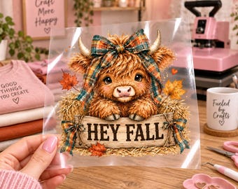 May include: A clear heat transfer decal featuring a fluffy brown Highland cow with a plaid bow between its horns, resting in hay. The cow holds a wooden sign that reads HEY FALL, decorated with orange autumn leaves and plaid bows.