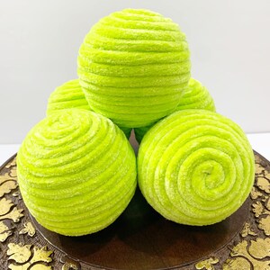 May include: Five lime green, round, decorative balls made of soft, fuzzy fabric. The balls are arranged in a pyramid shape on a brown wooden surface with gold accents.