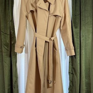 May include: A tan trench coat with a double-breasted design, a belt, and buttoned cuffs. The coat has a classic collar and epaulettes on the shoulders. The coat is hanging against a white background with green curtains on either side.