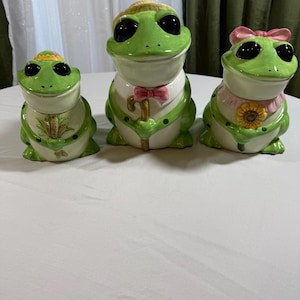 May include: Three ceramic frog figurines with green bodies, white bellies, and black eyes. The frogs wear hats, bow ties, and other accessories. The figurines are arranged on a white surface.