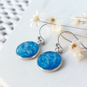 Op de afbeelding: Een paar ovale oorbellen met een helderblauwe, wervelende vormgeving. De oorbellen hebben een zilverkleurige metalen rand en haak. De achtergrond bevat witte bloemen en een wit oppervlak.
