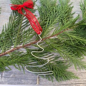 May include: A silver wire Christmas tree ornament with a red ribbon bow. The ornament is in the shape of a stylized tree and is displayed on a bed of green evergreen branches.