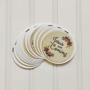 May include: A stack of round, white paper coasters with a beige circle in the center. The beige circle has a floral design and the text "Jesus isn't Coming" in black cursive lettering.