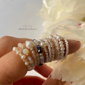 May include: A stack of delicate silver rings with white pearls, opals, and moonstone beads. The rings are worn on a finger against a white flower background. The text "VAIDA BluebellandClovers" is visible.