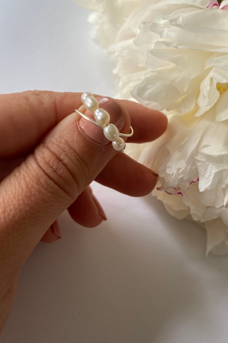 May include: A delicate silver ring with a row of white pearls. The ring is worn on a finger.