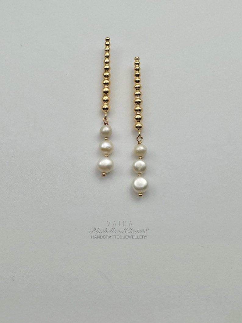 May include: A pair of gold-colored drop earrings. Each earring features a vertical strand of small, round beads, with three white, round pearls dangling below. The earrings are handcrafted jewelry.
