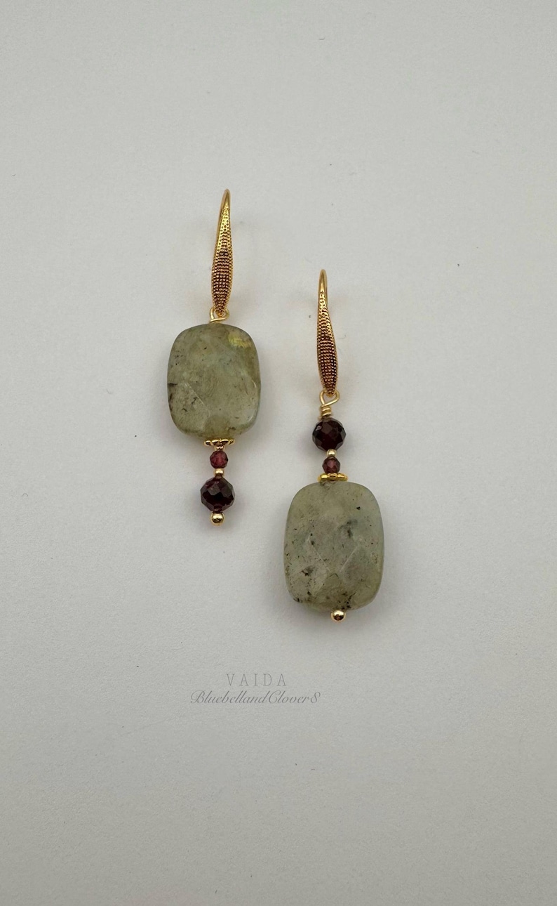 May include: A pair of gold earrings with a light green stone and dark red beads. The earrings are labeled "VAIDA BluebellandClover8".