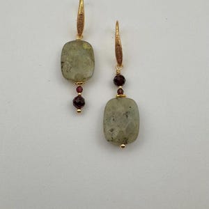 May include: A pair of gold earrings with a rectangular green stone and a small red bead. The earrings are hanging from a gold hook.